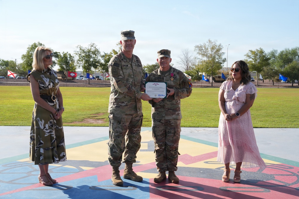 A military award ceremony is taking place outdoors on a painted parade ground, where a senior officer is presenting a certificate and medal to a lower-ranking soldier. Two civilian women, likely family members or guests, stand on either side of the two uniformed personnel. State and unit flags are visible in the background, suggesting a formal military installation setting.