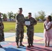 A military award ceremony is taking place outdoors on a painted parade ground, where a senior officer is presenting a certificate and medal to a lower-ranking soldier. Two civilian women, likely family members or guests, stand on either side of the two uniformed personnel. State and unit flags are visible in the background, suggesting a formal military installation setting.