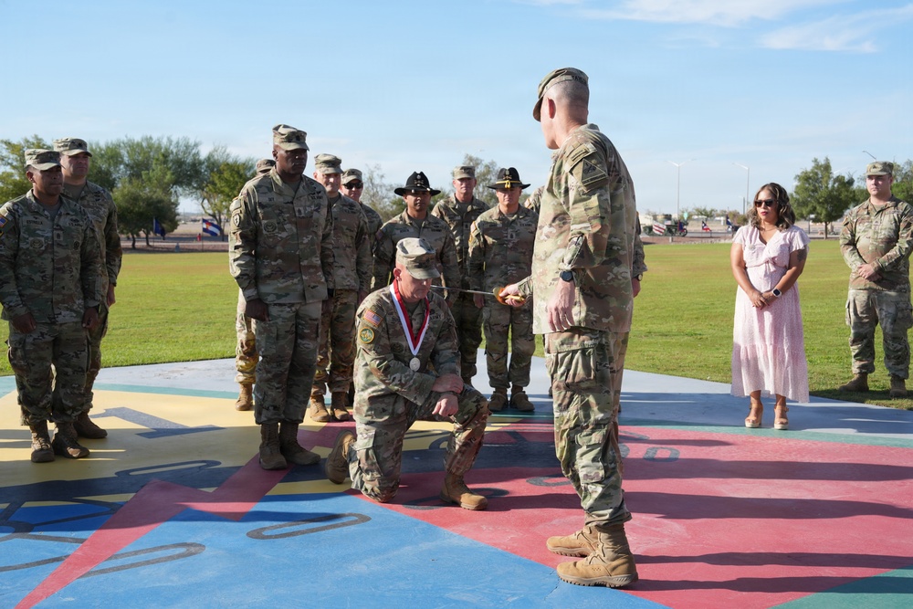 A military ceremony is taking place outdoors on a colorful painted surface, likely on a U.S. Army installation. A soldier is kneeling while receiving what appears to be a medal or award, being presented by a standing officer using a sword or saber. A formation of soldiers and one civilian woman in a pink dress observe the ceremony in the background.