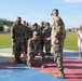 A military ceremony is taking place outdoors on a colorful painted surface, likely on a U.S. Army installation. A soldier is kneeling while receiving what appears to be a medal or award, being presented by a standing officer using a sword or saber. A formation of soldiers and one civilian woman in a pink dress observe the ceremony in the background.