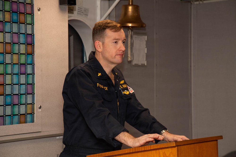A U.S. Navy personnel member in dark blue utility uniform stands at a wooden podium, appearing to speak or present. A cross insignia is visible on the collar, suggesting the individual may be a Navy chaplain. The setting appears to be aboard a naval vessel, indicated by the brass ship's bell and gray metal bulkheads in the background.