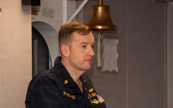 A U.S. Navy personnel member in dark blue utility uniform stands at a wooden podium, appearing to speak or present. A cross insignia is visible on the collar, suggesting the individual may be a Navy chaplain. The setting appears to be aboard a naval vessel, indicated by the brass ship's bell and gray metal bulkheads in the background.