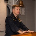 A U.S. Navy personnel member in dark blue utility uniform stands at a wooden podium, appearing to speak or present. A cross insignia is visible on the collar, suggesting the individual may be a Navy chaplain. The setting appears to be aboard a naval vessel, indicated by the brass ship's bell and gray metal bulkheads in the background.