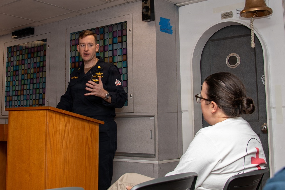 A U.S. Navy officer in dress blues is speaking at a wooden podium, appearing to address an audience. A person wearing a white shirt with a red cross emblem is seated in the foreground listening. The setting appears to be aboard a naval vessel, indicated by the ship's bell, watertight door, and porthole visible in the background.