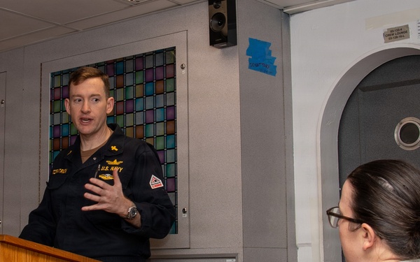 A U.S. Navy officer in dress blues is speaking at a wooden podium, appearing to address an audience. A person wearing a white shirt with a red cross emblem is seated in the foreground listening. The setting appears to be aboard a naval vessel, indicated by the ship's bell, watertight door, and porthole visible in the background.