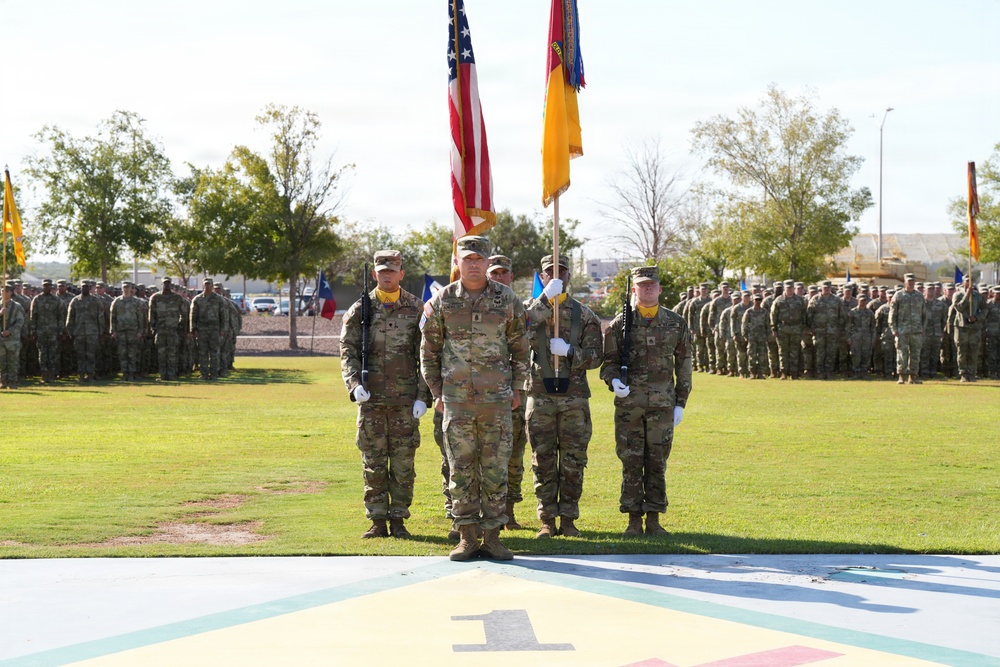 A formal U.S. Army military ceremony is taking place on a parade field, featuring a color guard presenting the American flag and unit guidons in the foreground. Large formations of soldiers in Army Combat Uniforms (ACU/OCP pattern) are standing at attention in the background. The painted ground markings and visible Texas state flag suggest this is likely Fort Bliss, Texas.