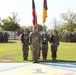A formal U.S. Army military ceremony is taking place on a parade field, featuring a color guard presenting the American flag and unit guidons in the foreground. Large formations of soldiers in Army Combat Uniforms (ACU/OCP pattern) are standing at attention in the background. The painted ground markings and visible Texas state flag suggest this is likely Fort Bliss, Texas.