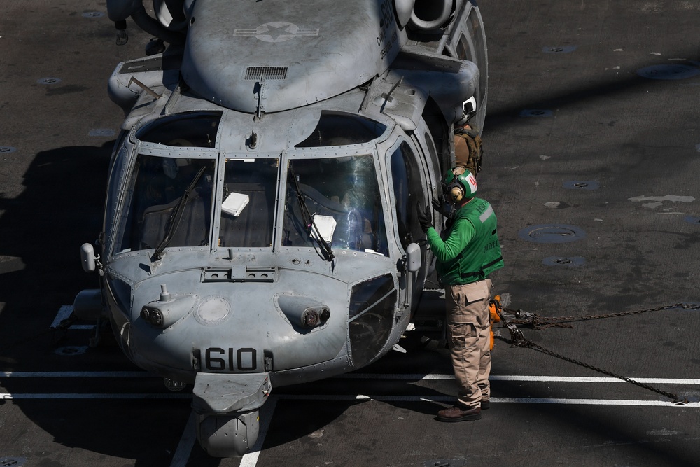 A military helicopter marked '610' is parked on what appears to be a naval aircraft carrier flight deck. A deck crew member wearing a green jersey and hearing protection is standing at the nose of the helicopter, likely performing a pre-flight check or communicating with the cockpit crew. The helicopter is secured to the deck with chains.