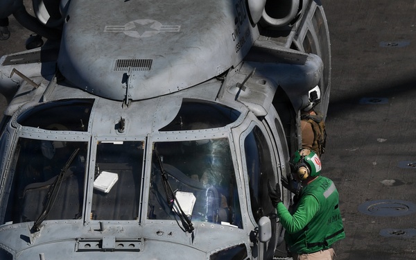 A military helicopter marked '610' is parked on what appears to be a naval aircraft carrier flight deck. A deck crew member wearing a green jersey and hearing protection is standing at the nose of the helicopter, likely performing a pre-flight check or communicating with the cockpit crew. The helicopter is secured to the deck with chains.