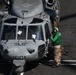 A military helicopter marked '610' is parked on what appears to be a naval aircraft carrier flight deck. A deck crew member wearing a green jersey and hearing protection is standing at the nose of the helicopter, likely performing a pre-flight check or communicating with the cockpit crew. The helicopter is secured to the deck with chains.