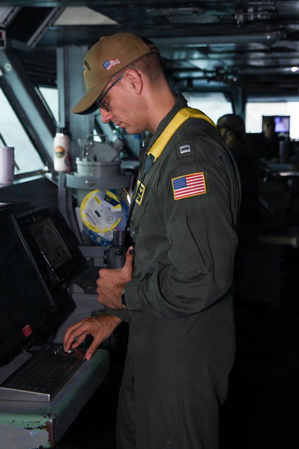 A U.S. Navy officer in a green flight suit is standing at a navigation console aboard what appears to be a naval vessel's bridge or combat information center. He is wearing a tan cap with a U.S. flag patch, glasses, a yellow inflatable life vest collar, and holds binoculars in one hand while operating a computer workstation with the other. Rank insignia and a U.S. flag patch are visible on his sleeve.