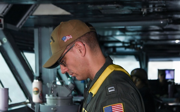 A U.S. Navy officer in a green flight suit is standing at a navigation console aboard what appears to be a naval vessel's bridge or combat information center. He is wearing a tan cap with a U.S. flag patch, glasses, a yellow inflatable life vest collar, and holds binoculars in one hand while operating a computer workstation with the other. Rank insignia and a U.S. flag patch are visible on his sleeve.