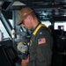 A U.S. Navy officer in a green flight suit is standing at a navigation console aboard what appears to be a naval vessel's bridge or combat information center. He is wearing a tan cap with a U.S. flag patch, glasses, a yellow inflatable life vest collar, and holds binoculars in one hand while operating a computer workstation with the other. Rank insignia and a U.S. flag patch are visible on his sleeve.