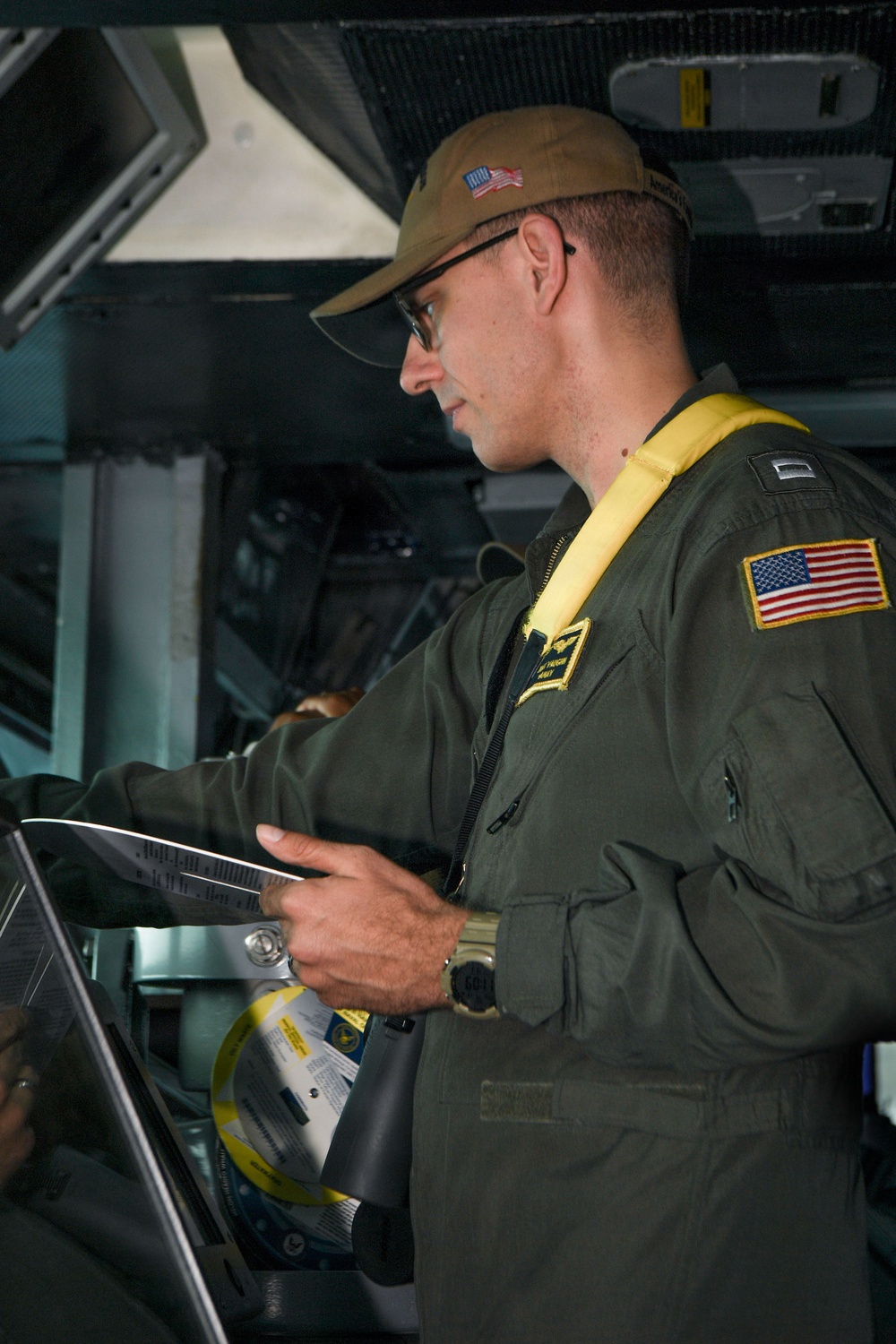 A U.S. military aviator in an olive green flight suit is reviewing documents or a checklist inside what appears to be a military aircraft. He is wearing a yellow inflatable life preserver vest, a tan cap with a U.S. flag patch, glasses, and a tactical watch. An American flag patch is visible on his right sleeve.