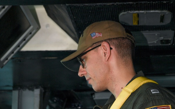 A U.S. military aviator in an olive green flight suit is reviewing documents or a checklist inside what appears to be a military aircraft. He is wearing a yellow inflatable life preserver vest, a tan cap with a U.S. flag patch, glasses, and a tactical watch. An American flag patch is visible on his right sleeve.