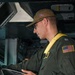 A U.S. military aviator in an olive green flight suit is reviewing documents or a checklist inside what appears to be a military aircraft. He is wearing a yellow inflatable life preserver vest, a tan cap with a U.S. flag patch, glasses, and a tactical watch. An American flag patch is visible on his right sleeve.