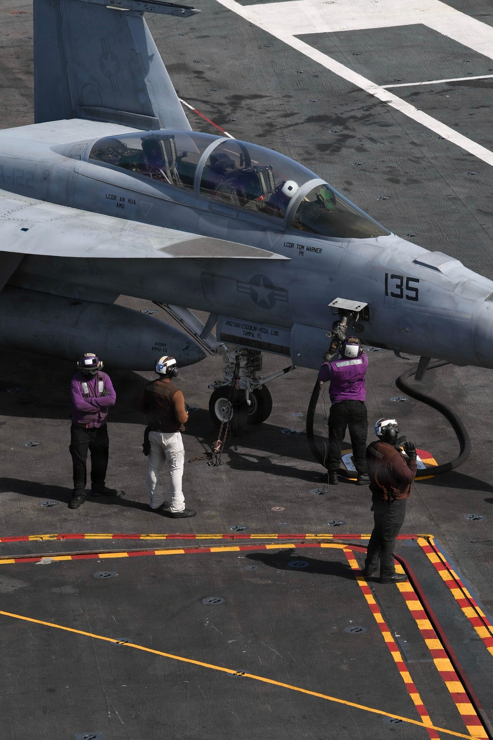 A U.S. Navy carrier-based jet aircraft numbered '135' is positioned on an aircraft carrier flight deck, with ground crew personnel in purple jerseys attending to it. The aircraft appears to be an F/A-18 Hornet or Super Hornet with two occupants visible in the cockpit. Ground crew are conducting pre-flight or fueling operations, with a fuel hose visible connected to the aircraft.