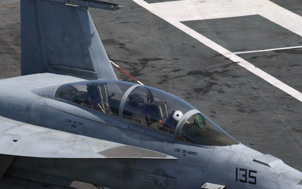A U.S. Navy carrier-based jet aircraft numbered '135' is positioned on an aircraft carrier flight deck, with ground crew personnel in purple jerseys attending to it. The aircraft appears to be an F/A-18 Hornet or Super Hornet with two occupants visible in the cockpit. Ground crew are conducting pre-flight or fueling operations, with a fuel hose visible connected to the aircraft.