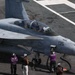 A U.S. Navy carrier-based jet aircraft numbered '135' is positioned on an aircraft carrier flight deck, with ground crew personnel in purple jerseys attending to it. The aircraft appears to be an F/A-18 Hornet or Super Hornet with two occupants visible in the cockpit. Ground crew are conducting pre-flight or fueling operations, with a fuel hose visible connected to the aircraft.