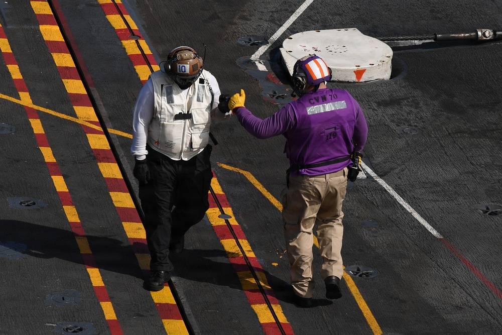Two naval personnel are standing on the flight deck of an aircraft carrier, appearing to communicate with hand signals or gestures. One individual wears a white vest with a brown helmet marked '10', while the other wears a purple shirt marked 'CVN 70' with a striped helmet. The deck features characteristic yellow and red striped markings typical of U.S. Navy carrier flight decks.