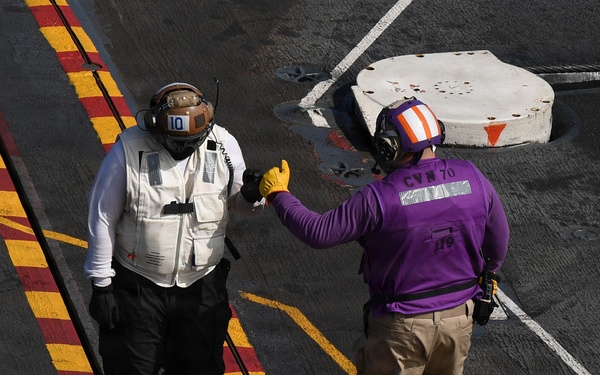 Two naval personnel are standing on the flight deck of an aircraft carrier, appearing to communicate with hand signals or gestures. One individual wears a white vest with a brown helmet marked '10', while the other wears a purple shirt marked 'CVN 70' with a striped helmet. The deck features characteristic yellow and red striped markings typical of U.S. Navy carrier flight decks.