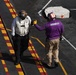 Two naval personnel are standing on the flight deck of an aircraft carrier, appearing to communicate with hand signals or gestures. One individual wears a white vest with a brown helmet marked '10', while the other wears a purple shirt marked 'CVN 70' with a striped helmet. The deck features characteristic yellow and red striped markings typical of U.S. Navy carrier flight decks.