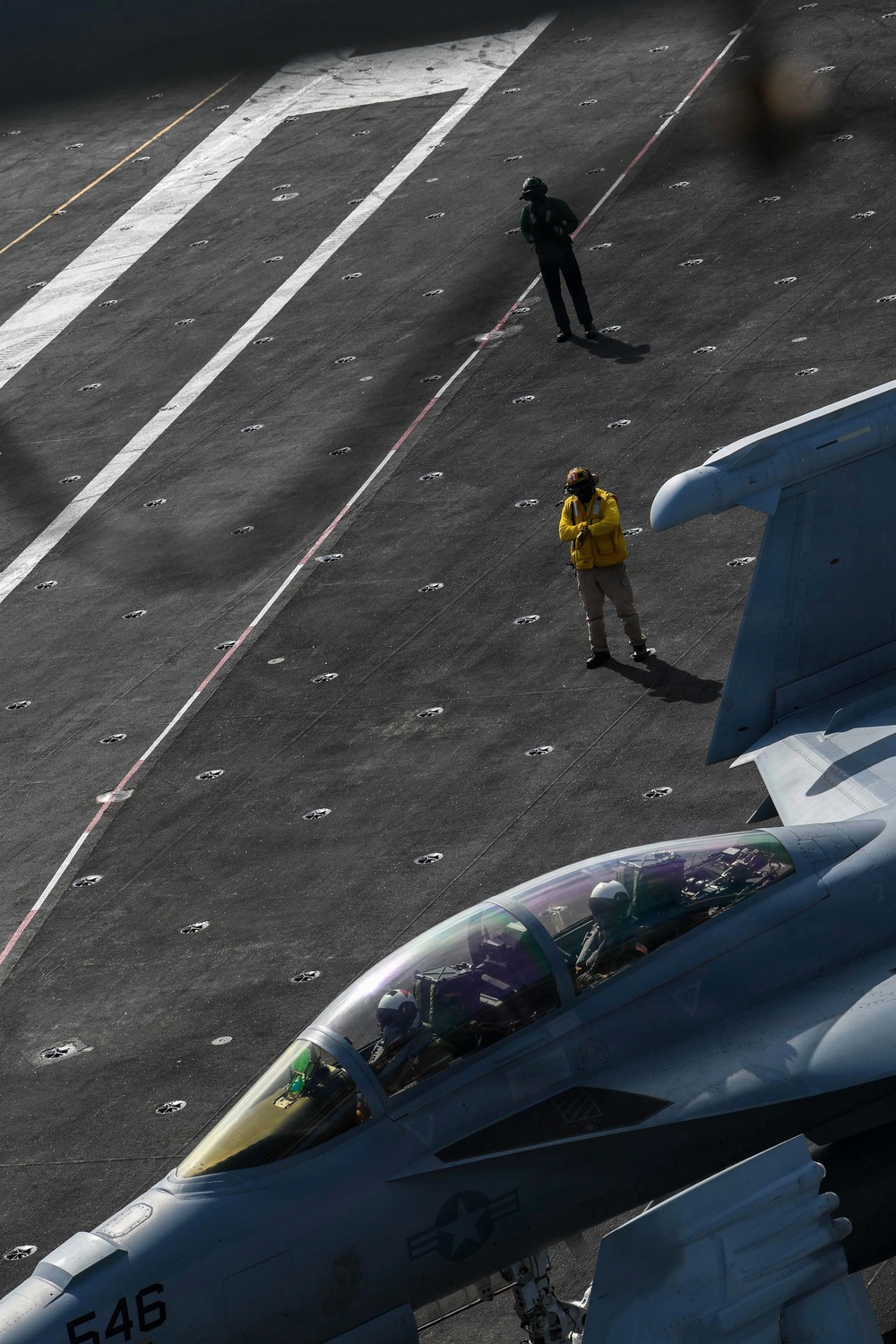 An aerial view of a naval aircraft carrier flight deck showing a two-seat fighter jet (tail number 546) in the foreground with at least one pilot visible in the cockpit. Two flight deck crew members are visible on the deck — one wearing a yellow vest and one in green — standing near the aircraft. The scene depicts pre-flight or taxi operations on an active carrier flight deck.
