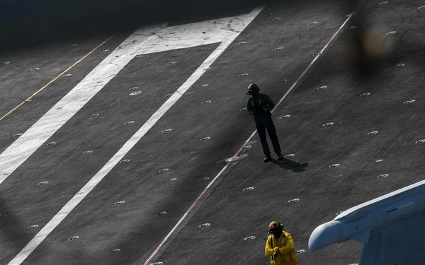 An aerial view of a naval aircraft carrier flight deck showing a two-seat fighter jet (tail number 546) in the foreground with at least one pilot visible in the cockpit. Two flight deck crew members are visible on the deck — one wearing a yellow vest and one in green — standing near the aircraft. The scene depicts pre-flight or taxi operations on an active carrier flight deck.