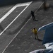 An aerial view of a naval aircraft carrier flight deck showing a two-seat fighter jet (tail number 546) in the foreground with at least one pilot visible in the cockpit. Two flight deck crew members are visible on the deck — one wearing a yellow vest and one in green — standing near the aircraft. The scene depicts pre-flight or taxi operations on an active carrier flight deck.