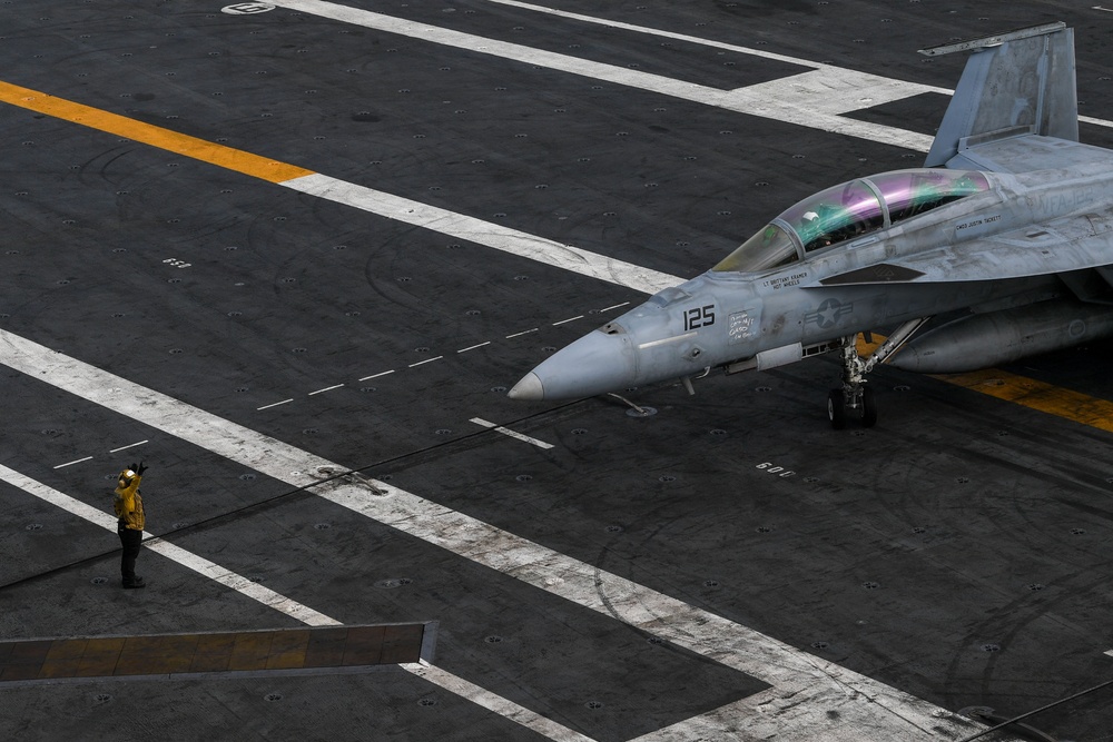 An aerial view of a U.S. Navy aircraft carrier flight deck showing a fighter jet numbered '125', identifiable as an F/A-18 Hornet or Super Hornet, positioned on the deck. A flight deck crew member wearing a yellow jersey is visible standing nearby, appearing to signal or direct the aircraft. The image captures the operational environment of a carrier flight deck with characteristic markings, catapult tracks, and deck equipment.