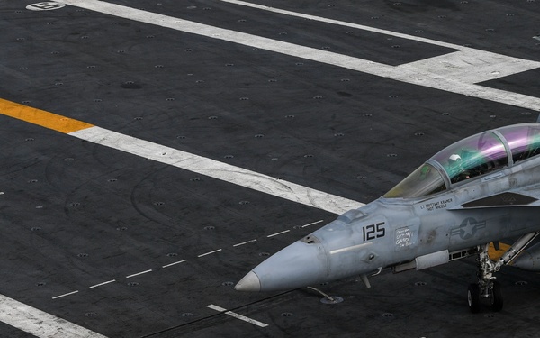An aerial view of a U.S. Navy aircraft carrier flight deck showing a fighter jet numbered '125', identifiable as an F/A-18 Hornet or Super Hornet, positioned on the deck. A flight deck crew member wearing a yellow jersey is visible standing nearby, appearing to signal or direct the aircraft. The image captures the operational environment of a carrier flight deck with characteristic markings, catapult tracks, and deck equipment.