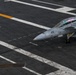 An aerial view of a U.S. Navy aircraft carrier flight deck showing a fighter jet numbered '125', identifiable as an F/A-18 Hornet or Super Hornet, positioned on the deck. A flight deck crew member wearing a yellow jersey is visible standing nearby, appearing to signal or direct the aircraft. The image captures the operational environment of a carrier flight deck with characteristic markings, catapult tracks, and deck equipment.