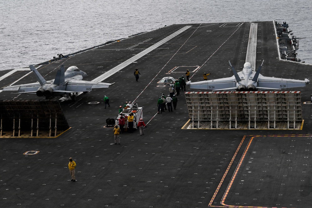 An aerial view of a large aircraft carrier flight deck at sea, showing at least two military jet aircraft positioned on the deck. Multiple crew members in color-coded jerseys are visible performing various flight deck operations. The image captures the expansive dark non-skid surface of the carrier with painted markings, catapult tracks, and jet blast deflectors raised.