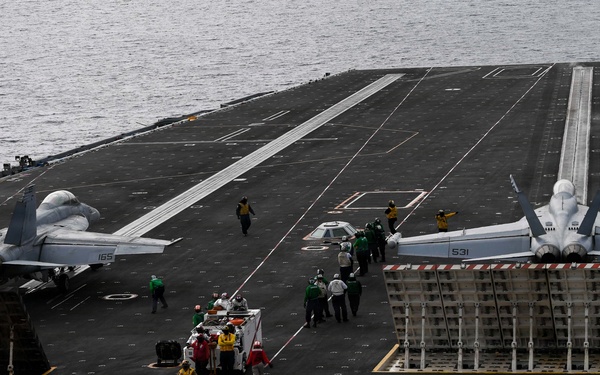 An aerial view of a large aircraft carrier flight deck at sea, showing at least two military jet aircraft positioned on the deck. Multiple crew members in color-coded jerseys are visible performing various flight deck operations. The image captures the expansive dark non-skid surface of the carrier with painted markings, catapult tracks, and jet blast deflectors raised.