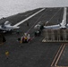 An aerial view of a large aircraft carrier flight deck at sea, showing at least two military jet aircraft positioned on the deck. Multiple crew members in color-coded jerseys are visible performing various flight deck operations. The image captures the expansive dark non-skid surface of the carrier with painted markings, catapult tracks, and jet blast deflectors raised.
