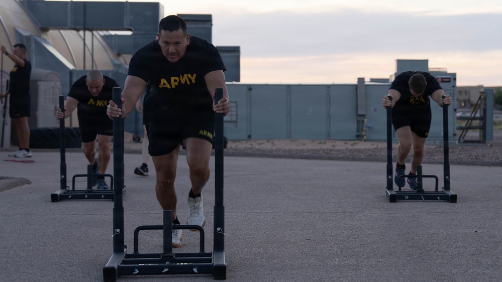 Three U.S. Army soldiers are performing sled push exercises on an outdoor paved surface, wearing matching black Army PT (Physical Training) uniforms. The exercise appears to be part of a structured physical fitness training session, likely the Army Combat Fitness Test (ACFT) or routine PT. The setting appears to be a military installation during early morning or evening hours based on the ambient lighting.