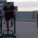 Three U.S. Army soldiers are performing sled push exercises on an outdoor paved surface, wearing matching black Army PT (Physical Training) uniforms. The exercise appears to be part of a structured physical fitness training session, likely the Army Combat Fitness Test (ACFT) or routine PT. The setting appears to be a military installation during early morning or evening hours based on the ambient lighting.