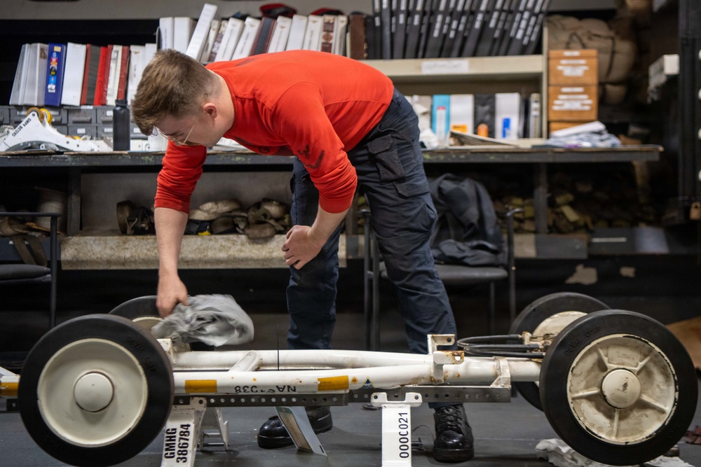 A young person wearing a red long-sleeve shirt and dark cargo pants is leaning over and working on what appears to be a lunar roving vehicle (LRV) chassis or similar wheeled frame artifact. The setting appears to be a museum storage or conservation workspace, with shelving units filled with binders, documents, and various artifacts visible in the background. The object being examined bears identification labels including 'GMHG 386784' and 'C021', suggesting it is a catalogued museum artifact.
