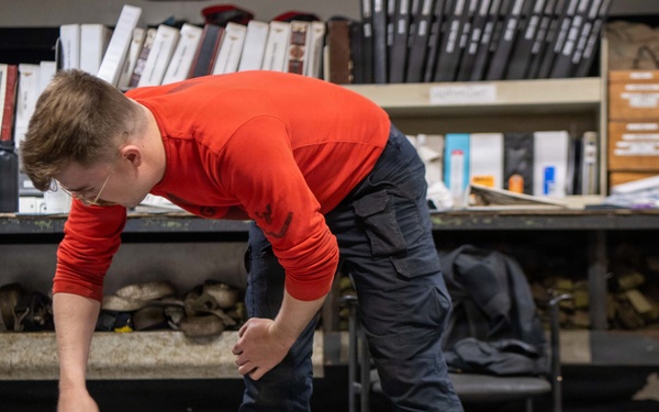 A young person wearing a red long-sleeve shirt and dark cargo pants is leaning over and working on what appears to be a lunar roving vehicle (LRV) chassis or similar wheeled frame artifact. The setting appears to be a museum storage or conservation workspace, with shelving units filled with binders, documents, and various artifacts visible in the background. The object being examined bears identification labels including 'GMHG 386784' and 'C021', suggesting it is a catalogued museum artifact.