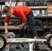 A young person wearing a red long-sleeve shirt and dark cargo pants is leaning over and working on what appears to be a lunar roving vehicle (LRV) chassis or similar wheeled frame artifact. The setting appears to be a museum storage or conservation workspace, with shelving units filled with binders, documents, and various artifacts visible in the background. The object being examined bears identification labels including 'GMHG 386784' and 'C021', suggesting it is a catalogued museum artifact.