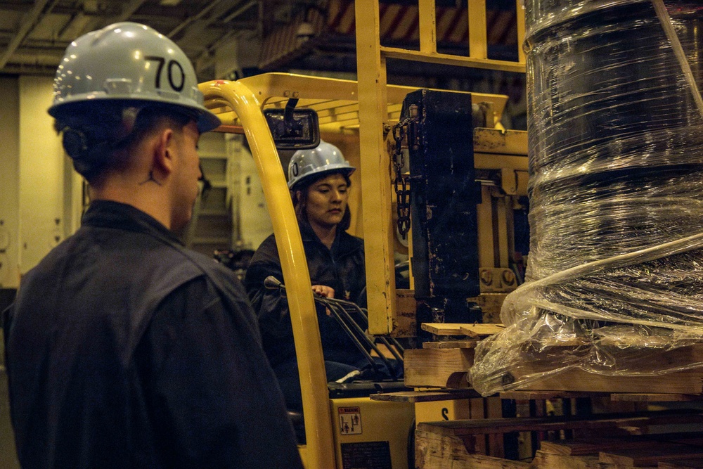 Two workers in hard hats and dark coveralls are operating a yellow forklift in what appears to be an industrial or naval vessel interior. One worker is seated at the forklift controls while the other stands nearby observing or directing the operation. The forklift is carrying a large spool or reel of cable wrapped in plastic sheeting on a wooden pallet.
