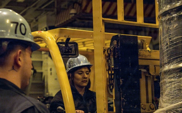Two workers in hard hats and dark coveralls are operating a yellow forklift in what appears to be an industrial or naval vessel interior. One worker is seated at the forklift controls while the other stands nearby observing or directing the operation. The forklift is carrying a large spool or reel of cable wrapped in plastic sheeting on a wooden pallet.