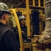 Two workers in hard hats and dark coveralls are operating a yellow forklift in what appears to be an industrial or naval vessel interior. One worker is seated at the forklift controls while the other stands nearby observing or directing the operation. The forklift is carrying a large spool or reel of cable wrapped in plastic sheeting on a wooden pallet.