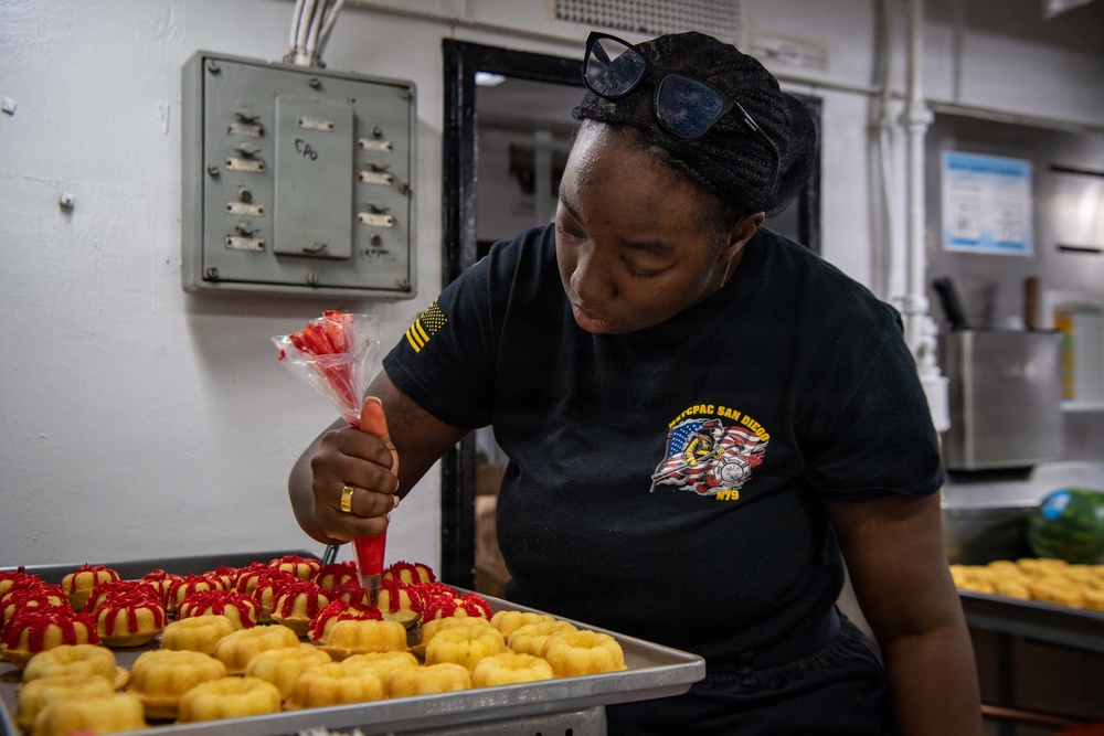 A person wearing a black military-style shirt with 'NTCPAC San Diego' insignia is decorating donuts with red icing using a piping bag in what appears to be a galley or institutional kitchen. The donuts are arranged on a metal baking tray, with some already decorated with red frosting patterns. The setting suggests a U.S. Navy ship or base food service environment.