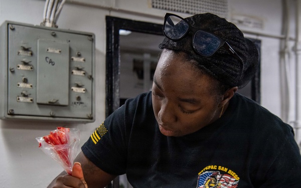 A person wearing a black military-style shirt with 'NTCPAC San Diego' insignia is decorating donuts with red icing using a piping bag in what appears to be a galley or institutional kitchen. The donuts are arranged on a metal baking tray, with some already decorated with red frosting patterns. The setting suggests a U.S. Navy ship or base food service environment.