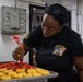 A person wearing a black military-style shirt with 'NTCPAC San Diego' insignia is decorating donuts with red icing using a piping bag in what appears to be a galley or institutional kitchen. The donuts are arranged on a metal baking tray, with some already decorated with red frosting patterns. The setting suggests a U.S. Navy ship or base food service environment.