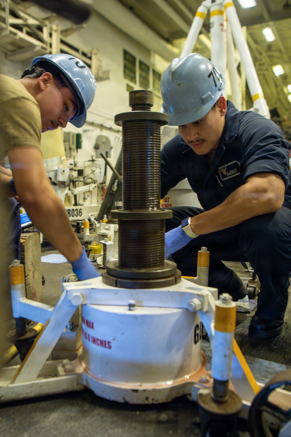Two workers wearing blue hard hats and blue nitrile gloves are collaborating on a mechanical maintenance task in what appears to be a naval vessel's hangar or machinery space. They are working on a large threaded spindle or jack screw assembly mounted on a white base unit with safety markings. The worker on the right is wearing a dark blue uniform consistent with U.S. Navy enlisted coveralls.