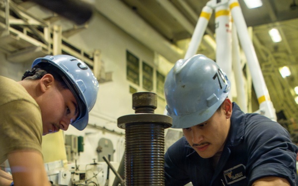 Two workers wearing blue hard hats and blue nitrile gloves are collaborating on a mechanical maintenance task in what appears to be a naval vessel's hangar or machinery space. They are working on a large threaded spindle or jack screw assembly mounted on a white base unit with safety markings. The worker on the right is wearing a dark blue uniform consistent with U.S. Navy enlisted coveralls.