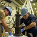 Two workers wearing blue hard hats and blue nitrile gloves are collaborating on a mechanical maintenance task in what appears to be a naval vessel's hangar or machinery space. They are working on a large threaded spindle or jack screw assembly mounted on a white base unit with safety markings. The worker on the right is wearing a dark blue uniform consistent with U.S. Navy enlisted coveralls.