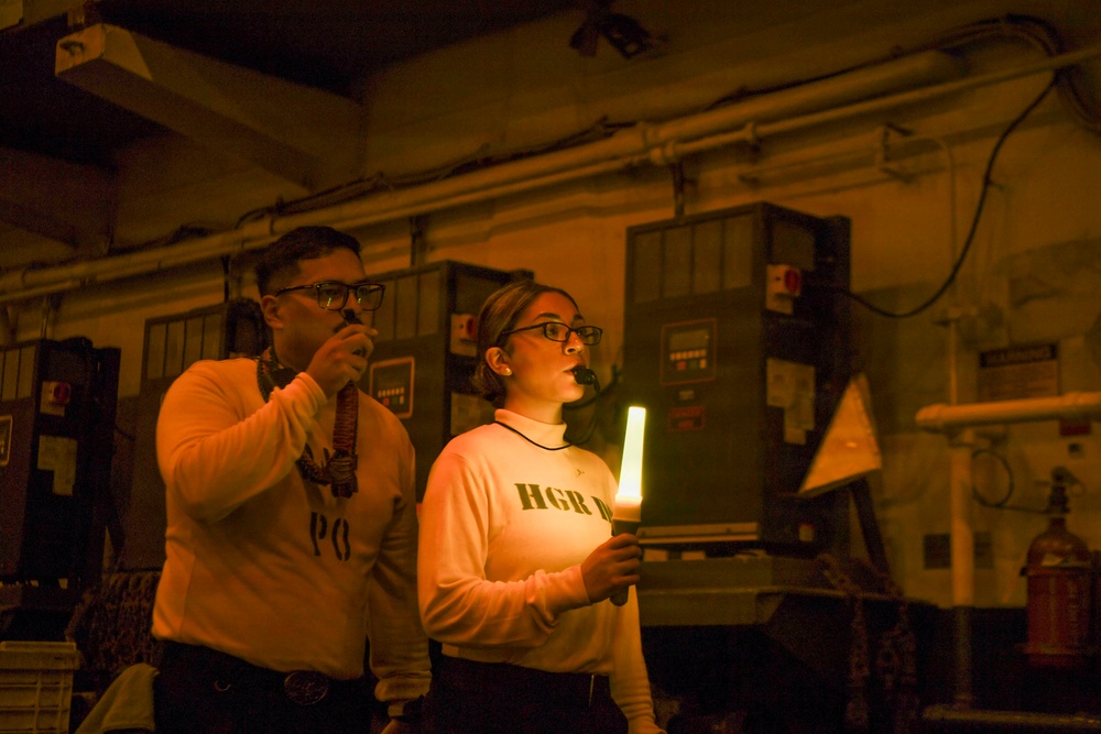 Two individuals are standing in a dimly lit industrial or mechanical space, illuminated primarily by warm orange emergency lighting and a glowing chemical light stick held by the woman on the right. Both are wearing matching sweatshirts with partial text visible ('PO' and 'HGR'). They appear to be conducting or participating in some kind of drill or operational exercise.